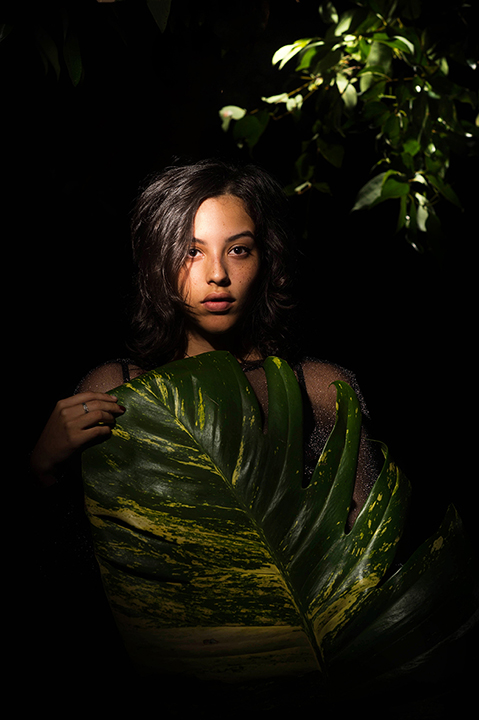 Fundo escuro com folhagem, mulher branca, cabelo curto castanho, semblante neutro, segurando uma grande planta Jiboia verde em frente ao corpo.
